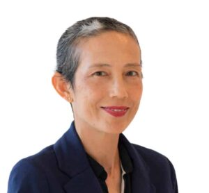 A person with short, graying hair and a warm smile is pictured against a white background. They are wearing a dark blue blazer and a black shirt.