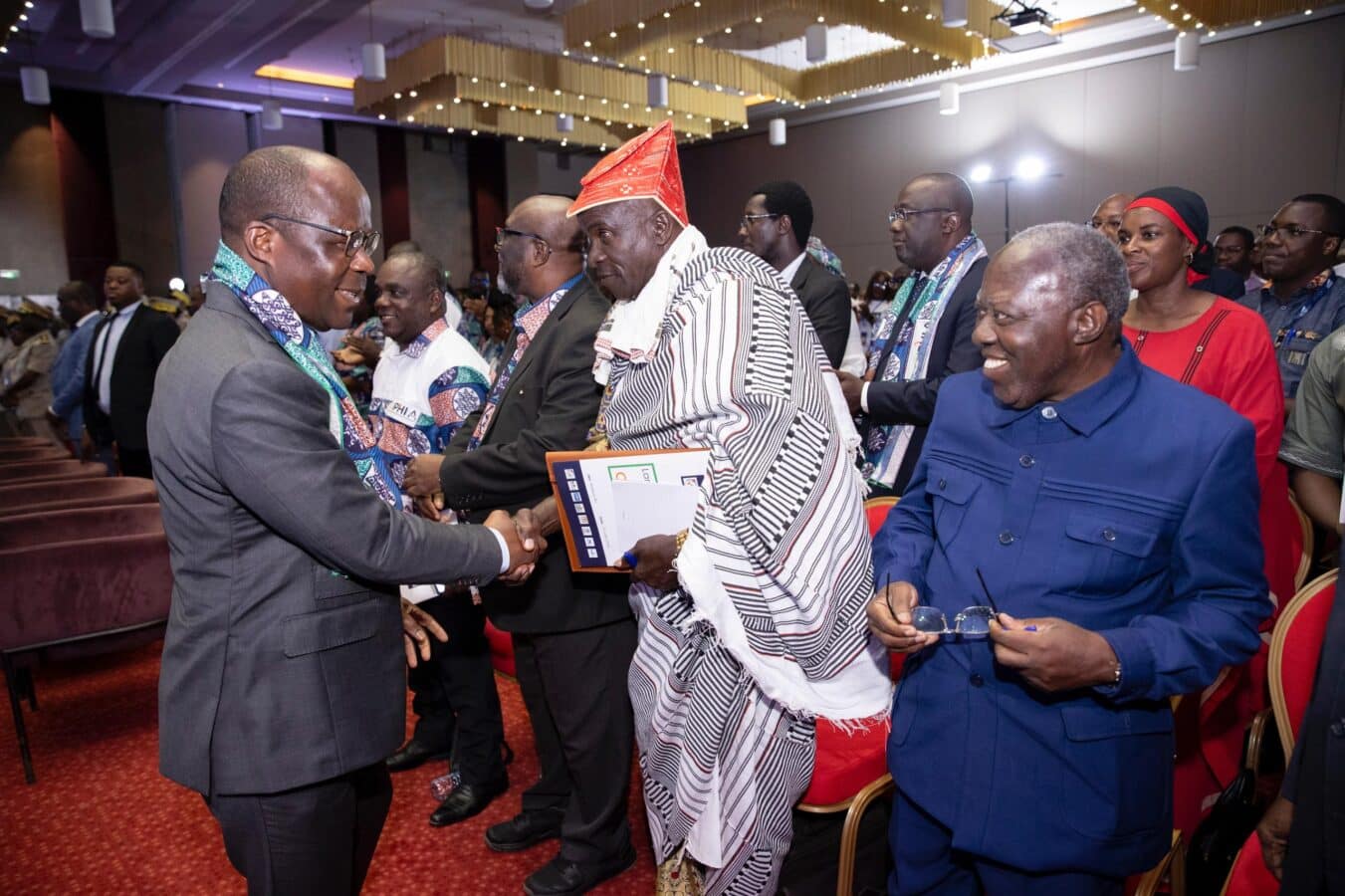 A group of people dressed in formal and traditional attire mingles in an indoor venue. Two individuals in the foreground exchange a handshake while others look on, smiling. The setting appears to be part of a larger event or ceremony, with red chairs and people gathered in the background.