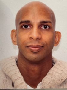 A person with a shaved head and large eyes, wearing a light-colored knitted sweater, looks directly at the camera against a plain background.