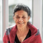 A woman with dark hair and a warm smile wears a black top and a red patterned shawl, standing in front of large windows with soft natural light.