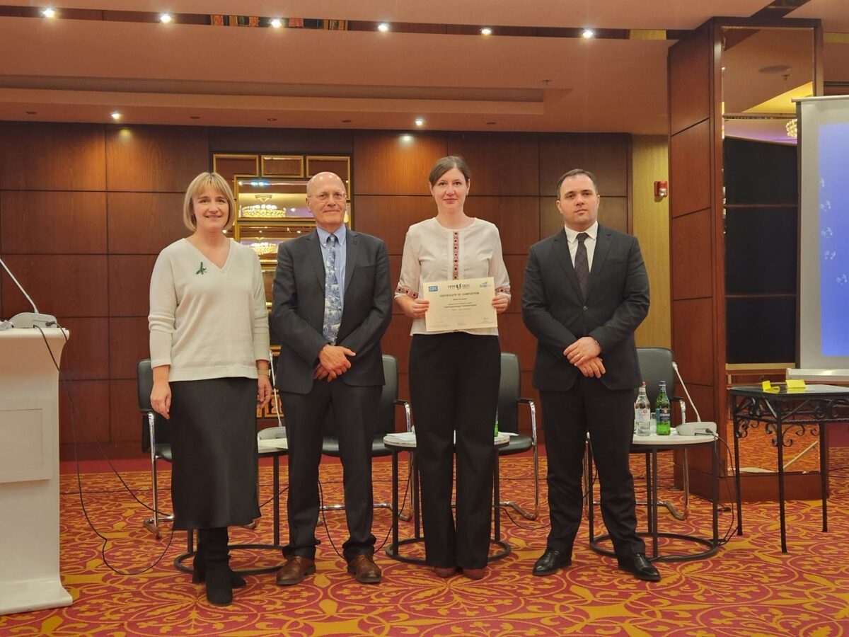 Four people pose for a photo in a conference room. The person second from the right holds a certificate. A podium is on the left, and chairs and a screen are in the background. They all appear formally dressed.