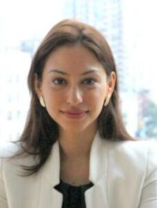 A person with long brown hair is smiling gently at the camera. They are wearing a white blazer over a black top, with large white earrings. The background is slightly blurred, showing an urban setting.