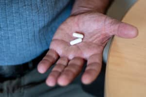 A close-up of an outstretched hand holding two white capsules, with part of a wooden table and a person wearing a blue shirt visible in the background.