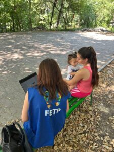 A woman in a blue vest labeled "FETP" works on a laptop while another woman, dressed in pink, sits on a green bench holding a toddler. They are outdoors, surrounded by trees and dry leaves.