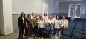 A group of twelve people, mostly women, stand together and smile in a room with blue chairs and arched windows. Most are wearing professional attire or white coats, suggesting a medical or academic setting.